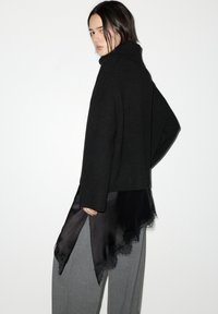 Schwarzer Strickpullover mit hohem Kragen, darüber ein schwarzes Satinstück mit Spitzenbesatz, kombiniert mit grauen, hoch taillierten Hosen.