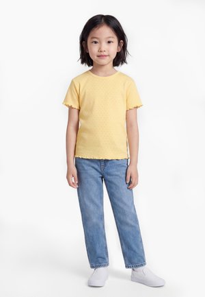 Ragazza giovane in piedi, indossa una maglietta gialla a maniche corte, jeans blu e scarpe bianche senza lacci, su uno sfondo bianco uniforme.