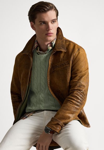 Brown leather jacket with a zip front and a single chest pocket. Layered with a green cable-knit sweater over a plaid shirt. White pants.