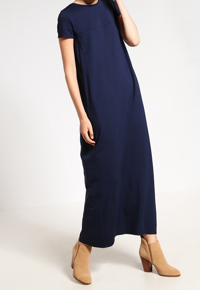 Femme portant une robe longue bleu marine à manches courtes et des bottines en daim couleur beige, debout devant un fond uni.