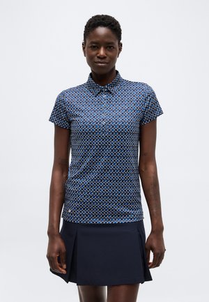 Žena má na sebe tmavomodrú polokošeľu s krátkym rukávom s vzorom a jednofarebnú tmavomodrú plisovanú sukňu, stojí pred jednofarebným bielym pozadím.