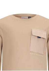 Beige Langarmhemd aus weichem Material, mit einer genähten Tasche auf der linken Brust, die marineblaue Akzente und eine strukturierte Druckdetail darunter aufweist.