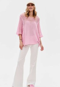 Roze, losgebreide, oversized trui met driekwartmouwen, gecombineerd met witte flared broeken en lichtroze schoenen. Pet op het hoofd.