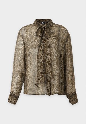 Femme debout portant un chemisier transparent à motif léopard avec un col à nœud, un pantalon noir slim et des chaussures slingback noires à talons hauts.