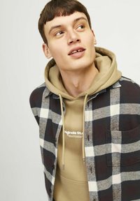 Rutede flanel skjorte i marineblå, grå og hvid over en beige hættetrøje med snore. Begge beklædningsgenstande viser et afslappet lagdelt design.