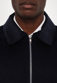 Navy corduroy jacket with a white collar, zip closure, and pronounced ribbed texture. Features a classic pointed collar design.