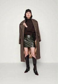 Casaco em padrão herringbone castanho, suéter castanho escuro, saia de couro verde, botas pretas até ao joelho com bico fino e um boné preto.