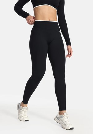 Persona che indossa leggings neri a vita alta con bordatura bianca e scarpe da ginnastica bianche, in piedi con una mano sul fianco e una gamba avanti.