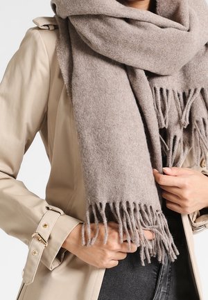 Persoon draagt een beige trenchcoat en wikkelt een grote taupekleurige wollen sjaal met franjes om hun nek en borst.