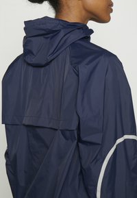 Marinblå vattentät jacka med huva, med en elegant, slät textur och reflexdetaljer längs ärmarna och fållen.