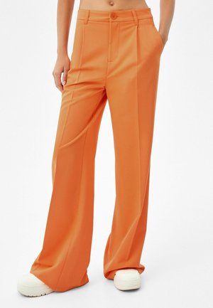 Person iført orange bukser med høj talje og vide ben, med hænderne i lommerne og hvide platformsneakers mod en hvid baggrund.