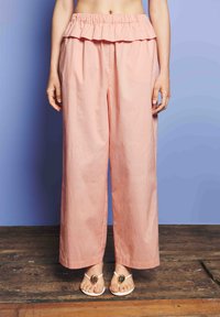 Nő magas derekú, laza rózsaszín nadrágban, fodros derekúval, fehér szandállal, arany ovális díszítéssel, fa padlón.