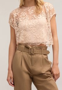Femme portant un crop top en dentelle rose transparent et un pantalon beige taille haute avec une ceinture assortie, se tenant devant un fond neutre.