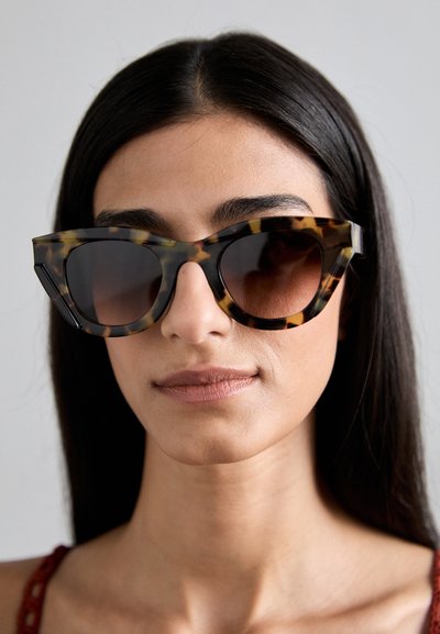 Tortoiseshell sunglasses with angular frame, dark lenses, and contrasting lighter patterns; reflective texture and oversized design.