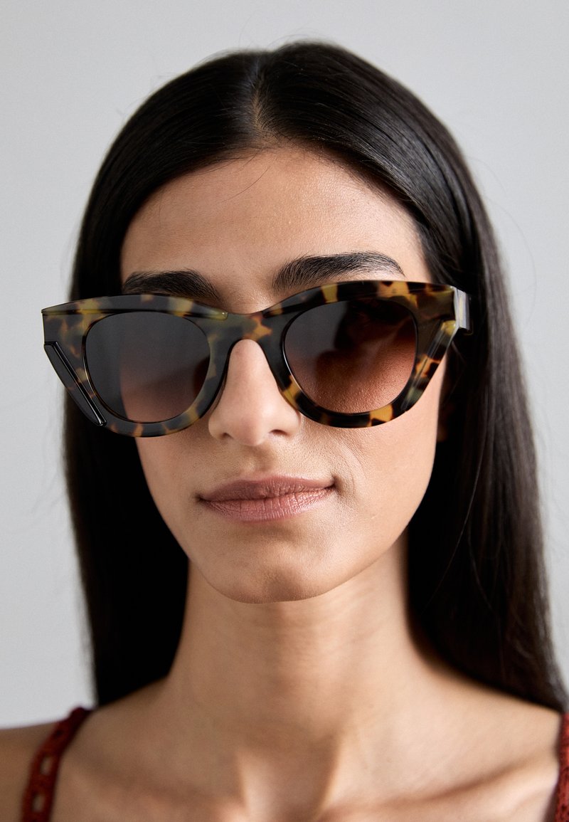 Tortoiseshell sunglasses with angular frame, dark lenses, and contrasting lighter patterns; reflective texture and oversized design.