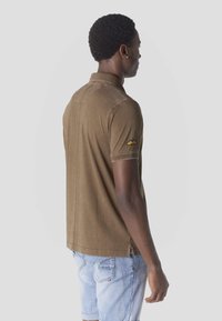 Moški, obrnjeni v stran, oblečen v rjavo polo majico s kratkimi rokavi z rumenim emblemom krila na rokavu in svetlo modre denim kratke hlače.