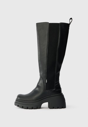 Crne čizme do koljena s debelim potplatom, elastičnim bočnim panelima, petljicom za povlačenje na vrhu i teksturiranom kožnom površinom na jednostavnoj pozadini.