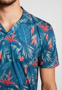 Camisa de manga corta en una tela azul con un patrón tropical que presenta hojas verdes y flores rojas. Diseño con cuello y textura suave.