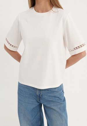 Vrouw draagt een wit T-shirt met korte mouwen en bloemrijke kanten randjes op de mouwen, en een blauwe spijkerbroek, staand met handen achter de rug.