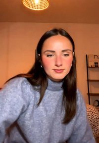 Ung kvinde med langt mørkt hår iført en lyseblå sweater, der sidder indendørs under en vævet lampeskærm med hylder og en plettet pude bag sig.