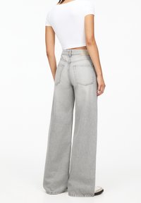 Donna vestita con jeans a gamba larga grigio chiaro e una maglietta bianca corta, in piedi con la schiena rivolta verso la fotocamera su uno sfondo bianco.