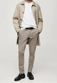 Béžový trenčkot přes bílé tričko, doplněný světle béžovými kalhotami a černými botami. Kabát má zapínání na knoflíky a boční rozparky.