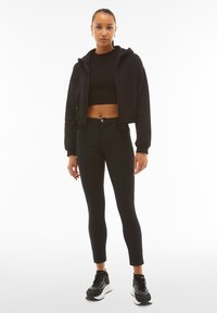 Mustat lyhyet hupparit, joissa on vetoketju, yhdistettyinä korkeavyötäröisiin mustiin farkkuihin. Mallilla on päällään mustat lenkkarit, joissa on teksturoitu pinta ja valkoinen pohja.