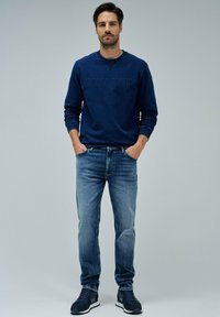 Mornarica dukserica s reljefnim logotipom "SALSA JEANS", uparena sa svijetloplavim farmerkama i tamnim tenisicama. Model stoji s rukama u džepovima.
