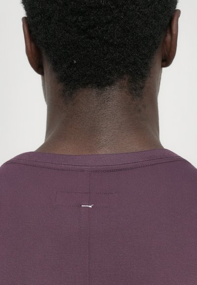 Back view of a person with short curly hair wearing a purple shirt with visible stitching at the neckline on a plain white background.