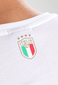 Camiseta de algodón blanca con un logo de la federación italiana de fútbol en oro y colores, acompañado de cuatro estrellas sobre el escudo.