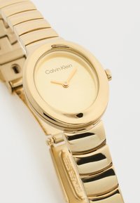 Ρολόι χειρός Calvin Klein σε χρυσούς τόνους με οβάλ καντράν, μίνιμαλ επιλογέα χωρίς αριθμούς και συνδεδεμένο μεταλλικό μπρασελέ σε λευκό φόντο.