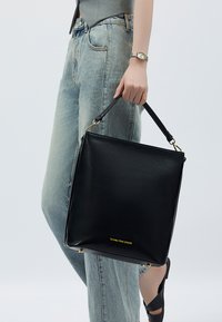 Borsa a mano in pelle nera con forma quadrata, hardware dorato e logo. La borsa è dotata di un manico corto e una texture liscia.