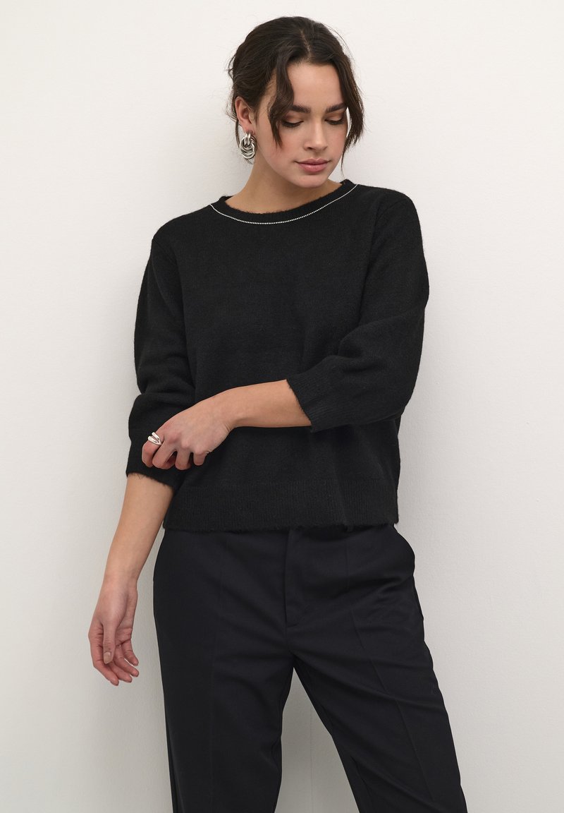 Pull noir texturé avec un col bateau, des manches trois-quarts et un ourlet brut. Associé à un pantalon foncé et des boucles d'oreilles en argent.