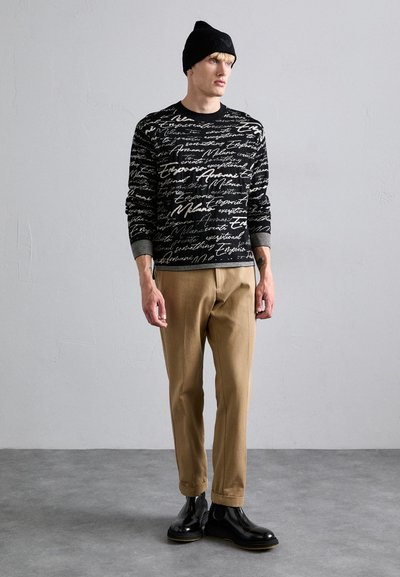 Schwarzer Pullover mit weißem handschriftlichem Textmuster, beigen Hosen und schwarzen Stiefeln. Das Model trägt eine schwarze Mütze. Einfaches, lässiges Outfit.