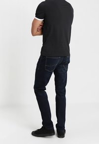 Camisa negra de manga corta con detalles en blanco, combinada con unos jeans de mezclilla azul oscuro. El modelo lleva zapatillas negras y se ve desde atrás.