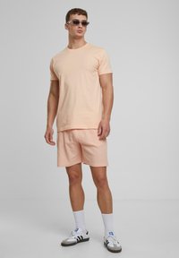Licht perzikkleurig katoen t-shirt en bijpassende geribbelde shorts. Model draagt een zonnebril, witte crew sokken en zwart-witte sneakers.