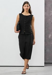 Black sleeveless top with textured fabric and button closure, paired with loose-fitting black cropped pants and black sandals. Carrying a small bag.