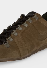 Chaussure en daim marron avec des lacets noirs passés à travers des crochets métalliques argentés et une semelle texturée visible au bord inférieur.