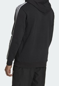 Svart huvtröja med tre vita ränder nedför axlarna och ärmarna, med ribbad nederkant och cuffs. Mjuk textur.