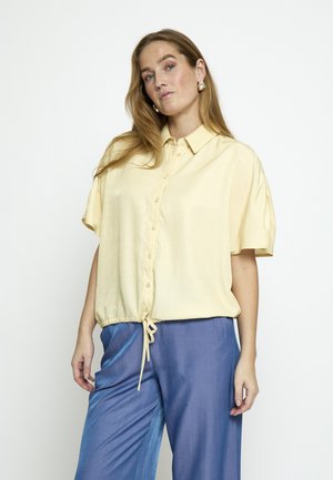 Žena s dugom svijetlosmeđom kosom, odjevena u blijedožutu košulju kratkih rukava s gumbima i široke plave hlače, stoji ispred bijele pozadine.