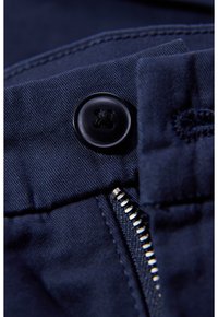 Mörk marinblått tyg på nära håll som visar en svart knapp, metallblixtlås och synliga sömnadsmönster. Texturerat material med slät yta.