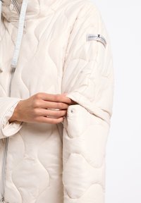 Beige gewatteerde jas met een golvend quiltpatroon, voorzien van een capuchon, trekkoord en een drukknoopsluiting bij de manchetten voor een verstelbare pasvorm.
