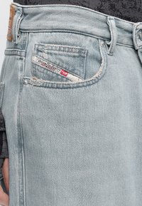 Φωτεινά μπλε τζιν από ντένιμ σε κοντινή λήψη που δείχνει μια μπροστινή τσέπη με ετικέτα Diesel Industry και θηλιές για ζώνη πάνω σε σκούρο τοπ.
