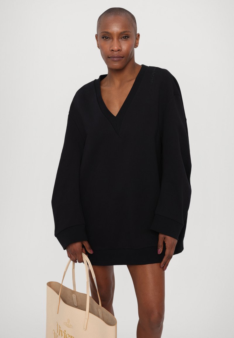Sweatshirt noir oversize avec un col en V, des manches longues et des poignets côtelés. Le modèle tient un sac fourre-tout beige avec des détails de logo en relief.