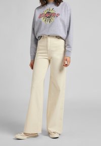 Persona che indossa pantaloni beige chiari a gamba larga, una felpa grigia con grafica del sole e la scritta "Kansas", e sneaker color avorio, in piedi davanti a uno sfondo neutro.