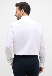 Camicia bianca a maniche lunghe con colletto standard, abbinata a pantaloni blu scuro con due tasche posteriori e dettagli con bottoni.