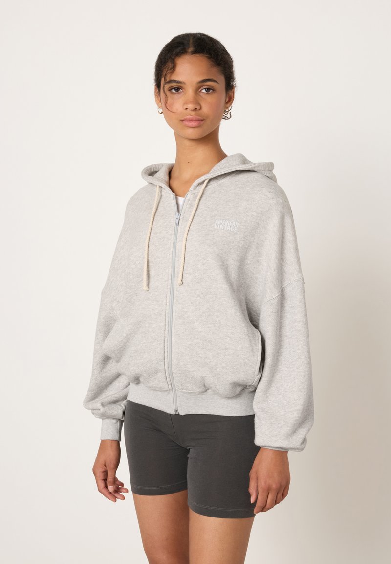 Siva jopa s kapuco, z zadrgo in majhnim logotipom. Ponaša se z elastičnim robom in manšetami, sproščenim krojem, nošena preko temnih hlač.