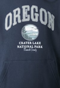 Námořnický modrý svetr s nápisem "OREGON" ve velkých krémových písmenech, s kruhovým grafickým prvkem jezera Crater a textem "Klamath County."