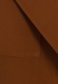 Brauner Stoff mit einer strukturierten Oberfläche. Verfügt über eine gefaltete Kante und eine kleine Taschenöffnung, sichtbare Nähte entlang der Kanten.