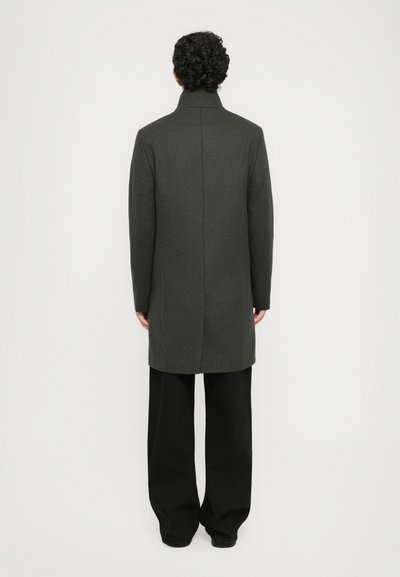 Manteau gris foncé avec un col haut, coupe droite et fente au dos, associé à un pantalon noir ample. Texture lisse, minimalisme dans les détails.